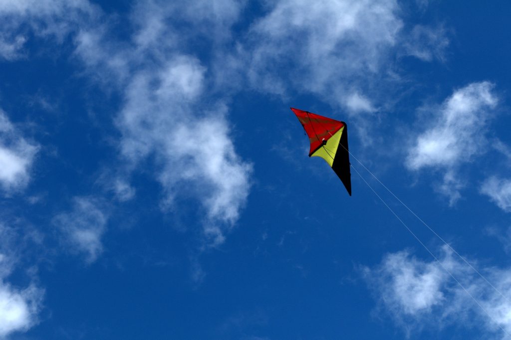 A black, red, and yellow kite flying high against a blue and white cloud sky.
