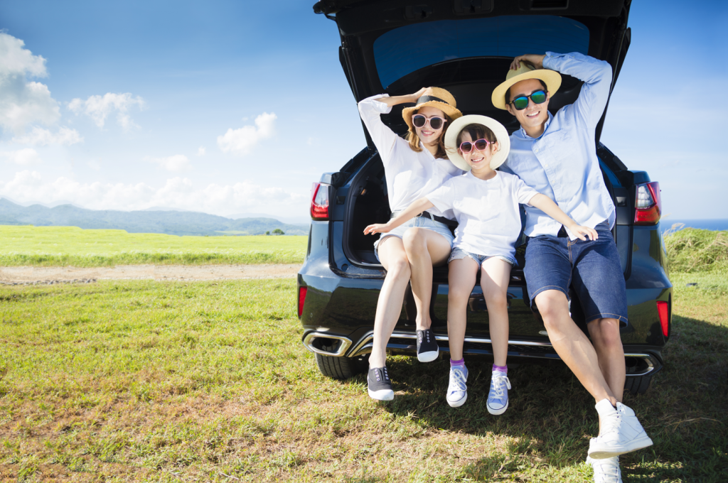 A family of three, mom, dad, and child, with sunglasses and sunhats on are sitting/resting on the trunk of their SUV in a field.